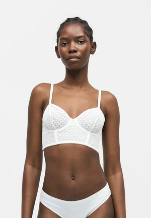 Γυναίκα που φοράει λευκό μακρύ σουτιέν από δαντέλα και απλά λευκά μπικίνι κάτω, στέκεται μπροστά σε απλό φωτεινό φόντο, κοιτάζοντας μπροστά.