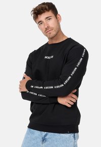 Sweatshirt noir à col rond, arborant "INDICODE" en blanc sur la poitrine et le long des manches. Texture douce et coupe décontractée.