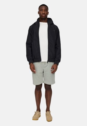 Schwarze leichte Jacke mit Kapuze, getragen über einem weißen Hemd und grauen Shorts. Das Modell steht in neutraler Haltung vor einem schlichten Hintergrund.