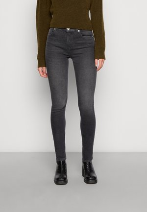 Kvinde iført sorte, højtaljede skinny jeans, sorte ankelstøvler med hæl og en olivengrøn langærmet trøje, stående på en grå gulv.