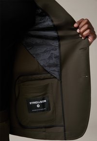 Olivengrünes Jackenfutter aus strukturiertem Gewebe, schwarze Akzenttasche mit "Strellson"-Logo. Das Futter zeigt ein dezentes Bergmuster.