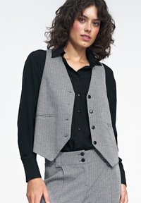 Szary i biały kamizelka w wzór jodełkowy z wyciętymi klapami, pięcioma guzikami i przednimi kieszeniami, nałożona na czarną koszulę z guzikiem.