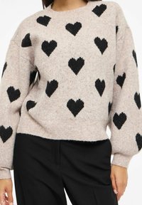 Světle růžový pletený svetr s černými vzory ve tvaru srdce, kulatým výstřihem a mírně zkráceným střihem. Texturovaná látka s balónovými rukávy.