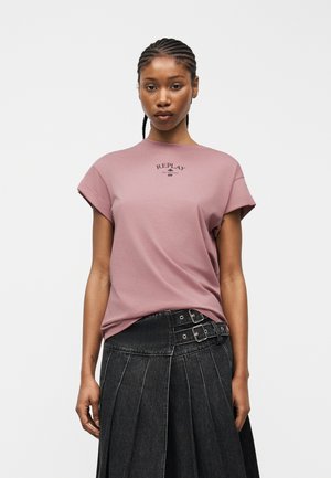 Donna con capelli intrecciati che indossa una t-shirt color malva con la scritta "Replay" e una gonna nera plissettata a vita alta con doppie fibbie, in piedi davanti a uno sfondo bianco.