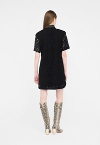 Kvinde iført en sort blonder kortærmet kjole og knæhøje støvler i slangeskind, stående med ryggen mod på en hvid baggrund.