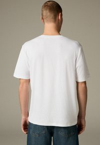 Camiseta blanca de algodón con cuello redondo y mangas cortas, que presenta una textura suave. La camiseta se lleva con jeans de mezclilla azul.