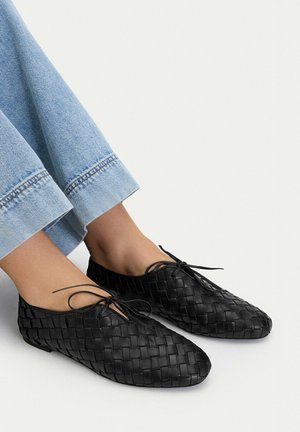Czarne plecione skórzane buty płaskie na sznurowadła, noszone z jasnoniebieskimi dżinsami, pokazane z boku na jednolitym tle.