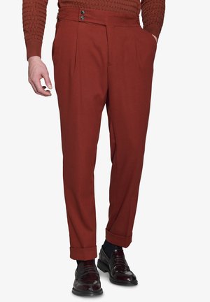 Pantaloni rosso ruggine realizzati in un tessuto liscio, con doppie pieghe, tasche laterali e bottoni neri in evidenza in vita. Orli con risvolto.