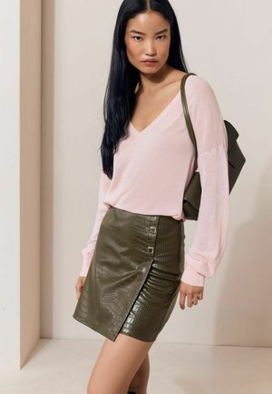 Mujer con cabello largo y negro lleva un suéter de cuello en V rosa claro, una falda de cuero texturizado verde oliva y lleva una mochila verde oliva a juego.