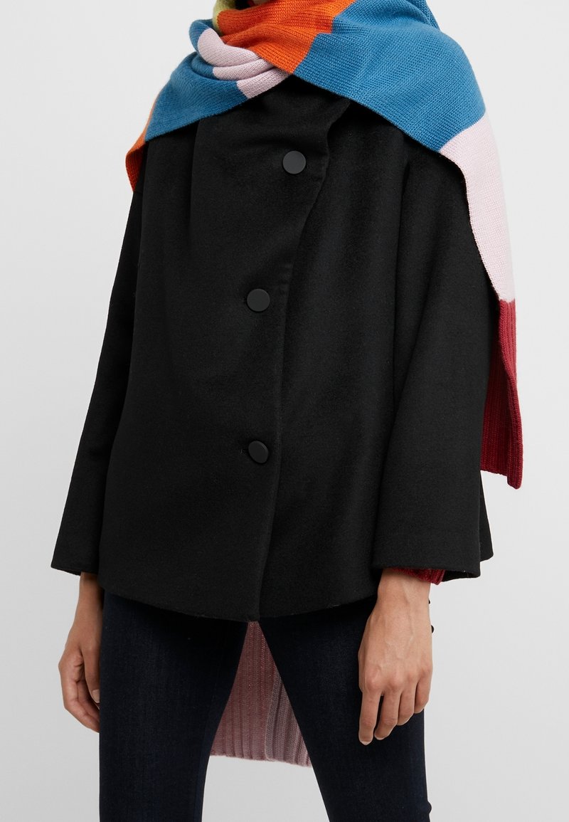 Personne portant un manteau noir avec de gros boutons et une écharpe en tricot multicolore drapée sur les épaules, visible du cou jusqu'aux hanches.