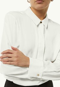 Camicia bianca a maniche lunghe con colletto button-down, texture liscia e dettagli in ottone dorato sui polsini e sul davanti.