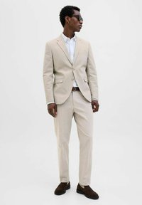 Beige suit with textured fabric, single-breasted jacket, and slim-fit trousers. White shirt underneath, brown shoes, and sunglasses.