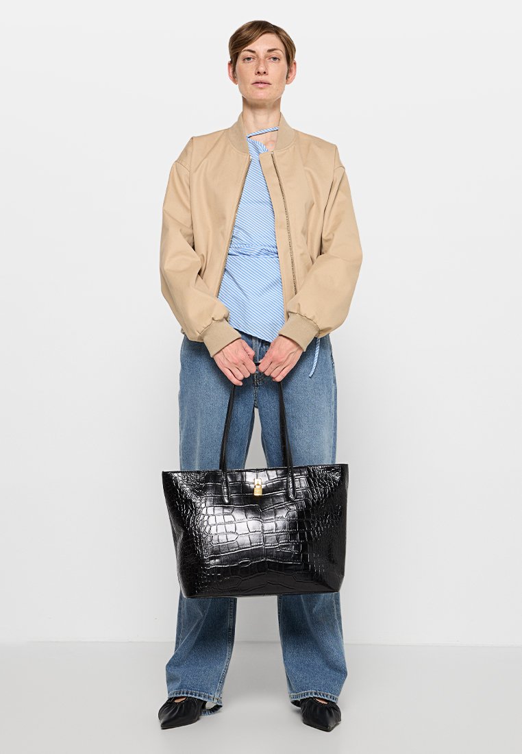 Black crocodile-patterned tote bag with two handles, shiny texture, and a gold hardware accent. Worn by a person in casual attire.