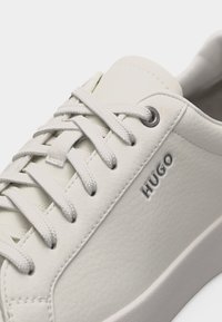 Λευκό δερμάτινο αθλητικό παπούτσι με λευκά κορδόνια, μεταλλικές τρύπες για κορδόνια, υφασμένο πέλμα και λογότυπο "HUGO" ανάγλυφο στην πλαϊνή επιφάνεια.