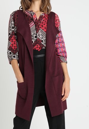 Vrouw draagt een blouse met rood, roze en zwart patroon met driekwart mouwen, een mouwloos lang bordeauxkleurig vest met zakken en een zwarte broek.