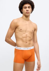 Πορτοκαλί βαμβακερά ανδρικά μποξεράκια με λευκή ελαστική ζώνη μέσης που φέρει το κείμενο και το λογότυπο "TOMMY HILFIGER". Εφαρμοστή σχεδίαση με ομαλή υφή.