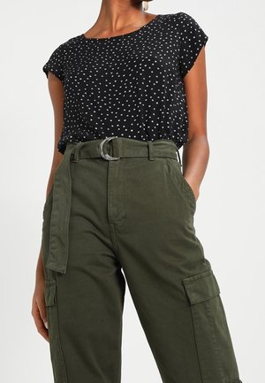 Femme portant un haut noir à manches courtes avec des taches blanches rentré dans un pantalon cargo taille haute vert olive avec une ceinture en tissu.