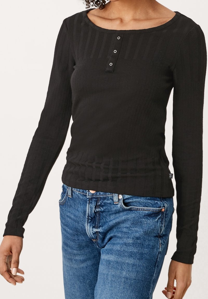 Person, der ein enges schwarzes Langarm-Rippoberteil mit drei silbernen Ösen am Ausschnitt und einer blauen Jeans trägt.
