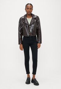 Ženska, oblečena v temno rjavo kratko usnjeno jakno, svetlo zeleno majico, črne oprijete hlače in črne loafer čevlje, stoji ob belem ozadju.