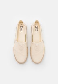 Zapatos slip-on de lona beige con bandas elásticas, superficie texturizada y detalles en encaje marrón claro en los bordes. Nombre de la marca "TOMS" visible en el interior.