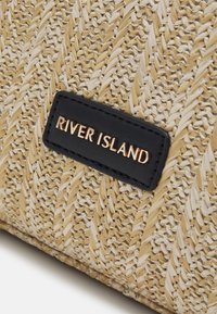 Woven straw bag with beige and cream textures. Features a black leather tag with "RIVER ISLAND" embossed in gold letters.