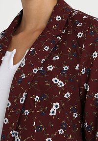 Marynarka w kwiaty w kolorze bordowym z białymi i niebieskimi kwiatami. Posiada wycięty kołnierz i miękką teksturę, zestawiona z białą koszulką z dekoltem w serek pod spodem.