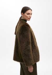 Brown faux fur coat with a loose fit, short sleeves, and a soft texture. Worn over a dark green top, with a plain background.