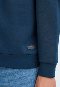 Sudadera navy con tejido texturizado, puños y dobladillo acanalados, que presenta una pequeña etiqueta negra con el nombre de la marca en el dobladillo lateral.