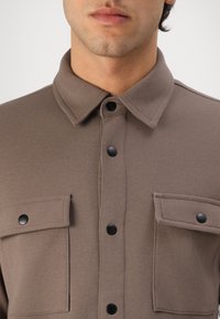 Camisa abotoada castanha, texturada, com colarinho pontudo, dois bolsos no peito e botões de pressão pretos para fecho.