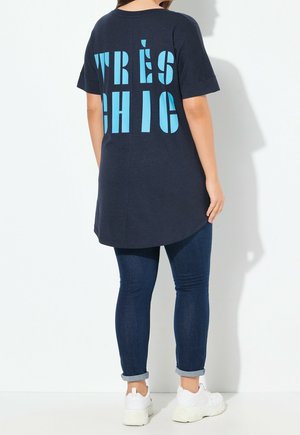 T-shirt oversize bleu marine avec ourlet arrondi, arborant un texte turquoise audacieux "TRÈS CHIC" dans le dos. Associé à un jean en denim foncé et des baskets blanches.