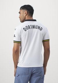 Bela nogometna majica z črnim ovratnikom in rokavi z rumenimi poudarki. "DORTMUND" je v črni barvi natisnjen na hrbtni strani. Material izgleda lahkoten in zračen.
