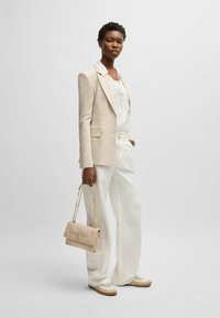 Blazer in lino beige sopra una maglietta bianca, abbinato a pantaloni larghi bianchi e sneakers beige. Tiene una borsa piccola beige con una tracolla dorata.