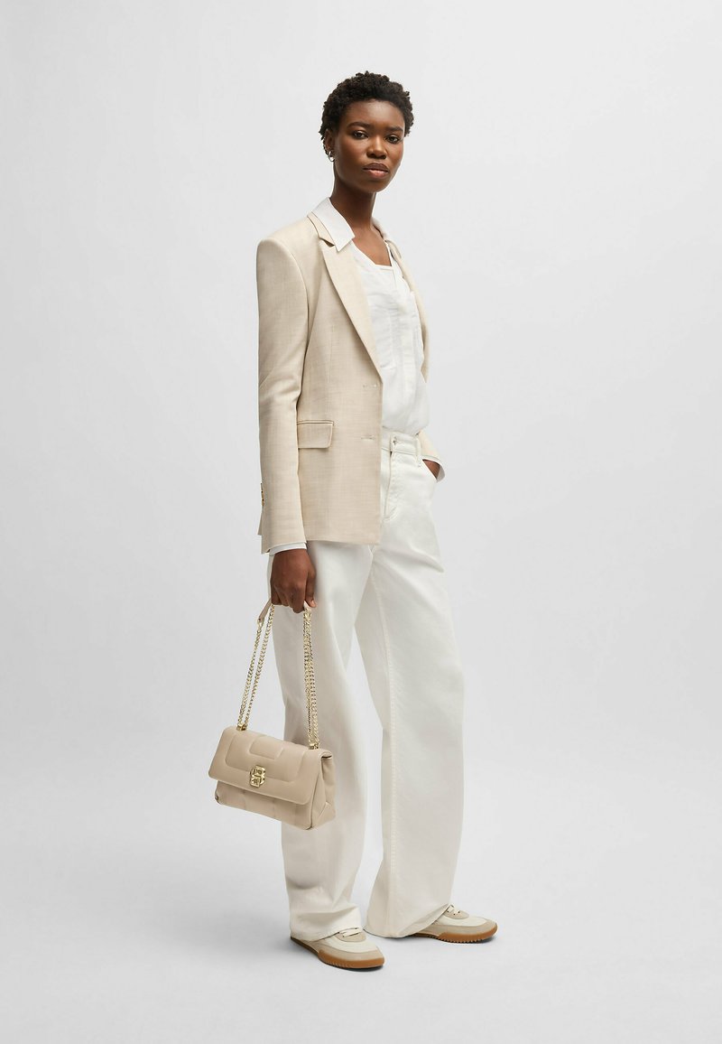 Blazer in lino beige sopra una maglietta bianca, abbinato a pantaloni larghi bianchi e sneakers beige. Tiene una borsa piccola beige con una tracolla dorata.