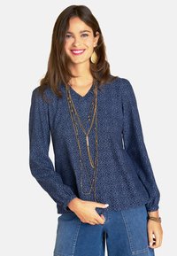 Marineblaue Bluse mit gemusterter Textur, V-Ausschnitt und langen Ärmeln. Kombiniert mit mehrlagigen goldenen Halsketten und Denim-Hosen.