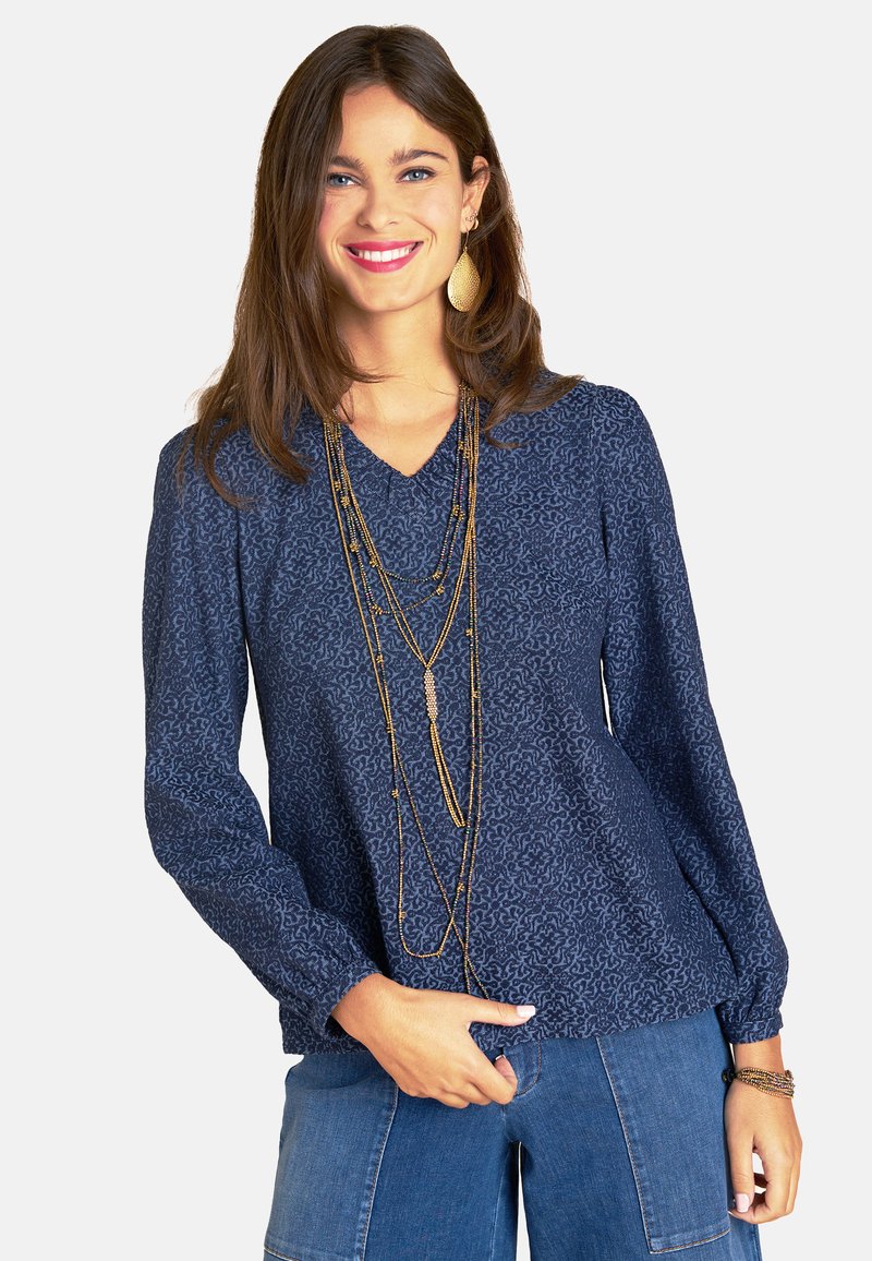 Marineblaue Bluse mit gemusterter Textur, V-Ausschnitt und langen Ärmeln. Kombiniert mit mehrlagigen goldenen Halsketten und Denim-Hosen.