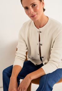 Ljust beige stickad cardigan med rund halsringning, långa ärmar och svarta knappar, i kombination med blå jeans, sittande på en trästol.