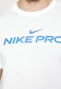 Vit Nike Pro t-shirt med korta ärmar, med en blå gradient "Nike Pro" logotyp och en liten blå swoosh-design. Mjuk textur.