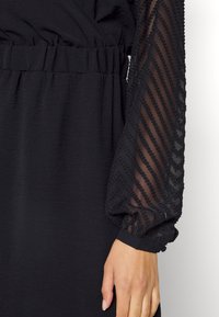 Robe noire avec taille élastique et manches longues transparentes, présentant des rayures diagonales texturées, portée par une personne avec une main visible.