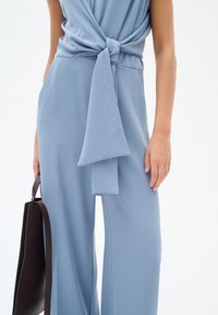 Salopette azzurra chiara con dettaglio in vita annodato, tessuto fluido, gambe larghe e texture liscia; accompagnata da una borsa a mano marrone scuro.