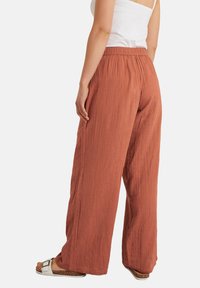 Terracotta broek met een losse pasvorm, gemaakt van een gestructureerde, gekreukte stof, voorzien van een elastische tailleband en wijde pijpen, gecombineerd met witte muiltjes.