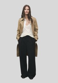 Trench coat beige sopra una maglietta bianca, abbinato a pantaloni neri a gamba larga. Il cappotto presenta un design doppiopetto e spalline sulle spalle.