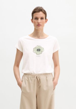 Vrouw met kort haar die een wit T-shirt met een groen bloemenpatroon en beige broek met trekkoord draagt, handen in de zakken, neutrale uitdrukking.