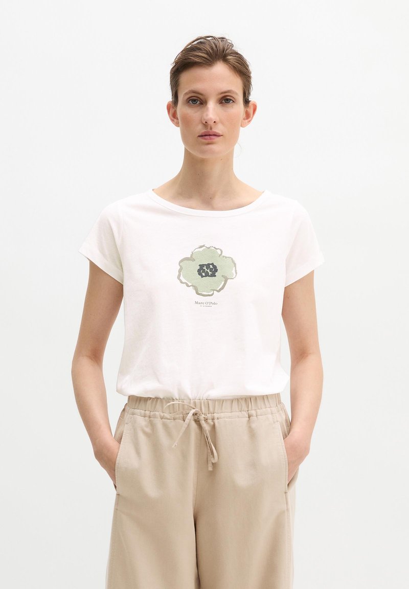 Femme aux cheveux courts portant un T-shirt blanc avec un imprimé de fleur verte et un pantalon beige à cordon, les mains dans les poches, expression neutre.