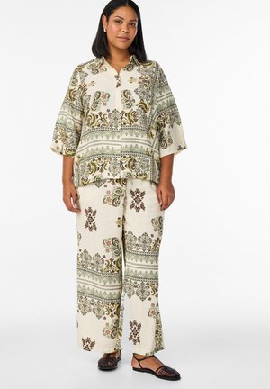 Žena má na sebe voľnú krémovú košeľu a široké nohavice so zelenými a hnedými kvetinovými a paisley vzormi, v kombinácii s čiernymi sandálmi.