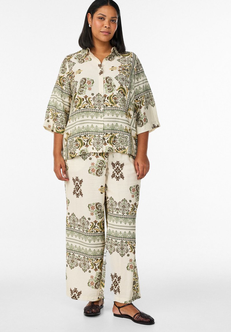 Mujer vistiendo una camisa cremosa holgada y pantalones anchos con patrones florales y de cachemira en verde y marrón, combinados con sandalias negras.