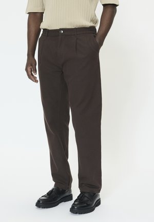 Brune bukser lavet af vævet stof, med en afslappet pasform, frontplisseringer og en enkelt knaplukning. Kombineret med sorte sko.