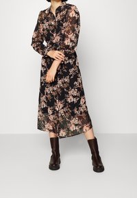 Kvinna i en svart genomsiktig midiklänning med rosa blommönster och långa ärmar, matchad med mörkbruna läderstövlar som går till vaden.