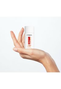 Fluide Revitalift Clinical de L'Oréal, bouteille cylindrique blanche avec du texte rouge, tenue dans une main sur un fond clair.