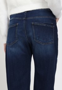 Donkerblauwe denim jeans met een gladde textuur, voorzien van twee achterzakken en stikseldetails. De taille heeft een klassieke snit.