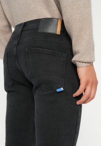 Jeans de denim negro con un corte ajustado, que presentan un parche de cuero en la cintura y una pequeña etiqueta azul en el bolsillo trasero.